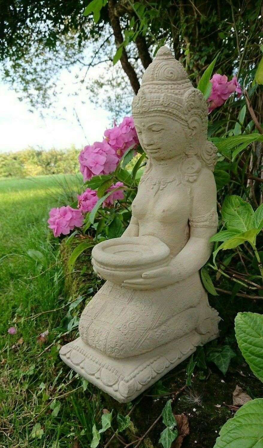 Stone Praying Geisha Garden Statue with intricate detailing and limestone finish, kneeling in a serene garden with pink flowers and greenery.