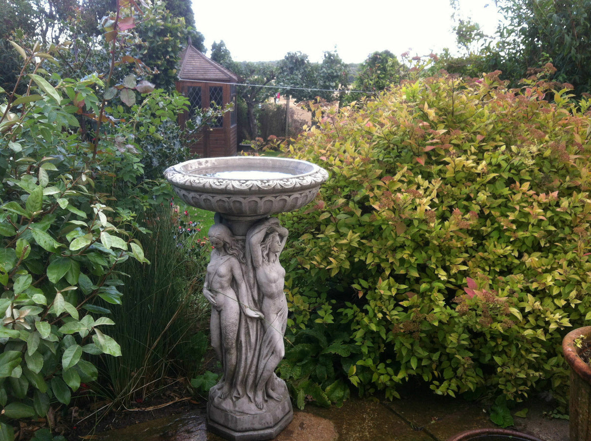 Three Graces Bird Bath in antique grey stone, standing tall in a vibrant garden with lush greenery and patio setting.