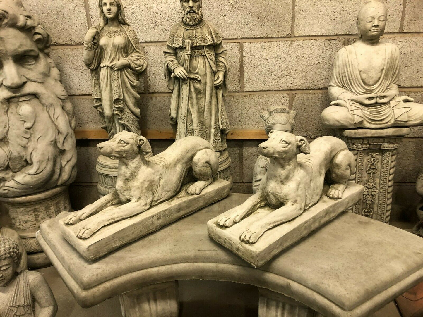 Pair of Stone Laying Greyhound Dog Statues displayed among classical garden sculptures, perfect for elegant garden settings.