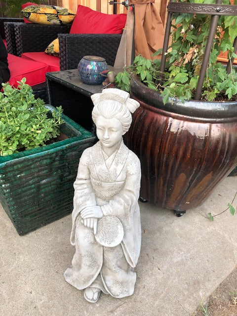 Stone Kneeling Geisha Lady Statue placed on patio among plants, enhancing outdoor decor with elegant Japanese-inspired design.