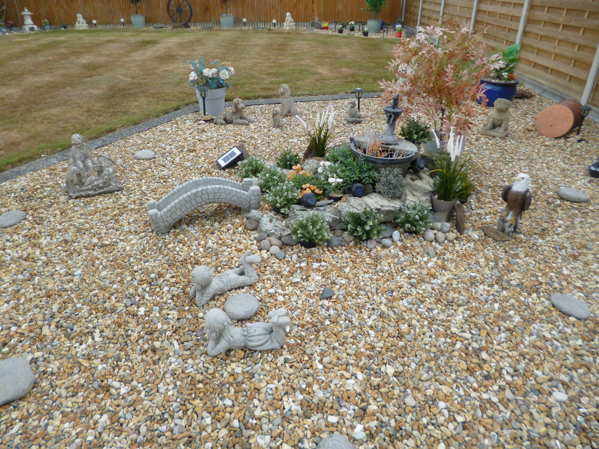 Garden ornament - stone pond bridge with antique finish in a landscaped pebble garden, surrounded by plants, statues, and solar lighting.