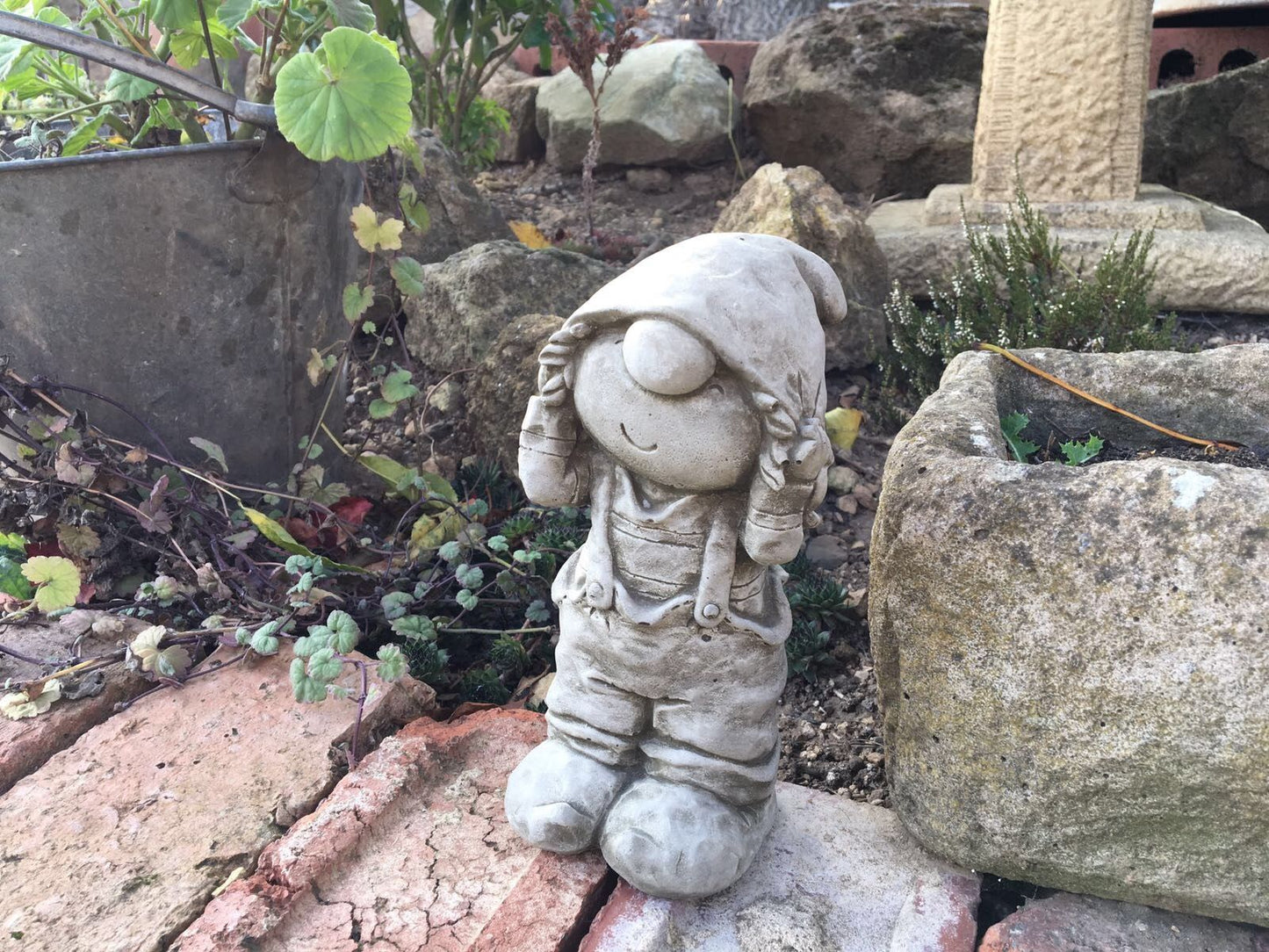 Stone Boy Garden Ornament in Dungarees standing on brickwork beside a planter. A delightful addition to garden paths or flowerbeds.