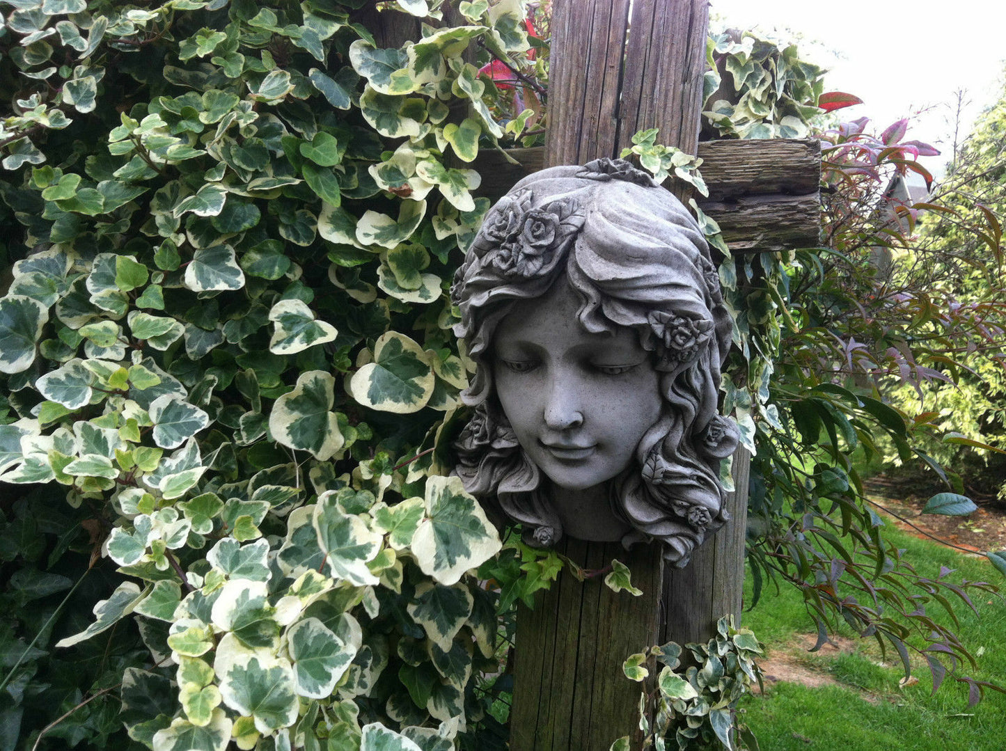 Lady Head Plaque Wall Art with floral crown mounted on wooden post, surrounded by ivy in garden. Antique stone finish adds charm to outdoor decor.