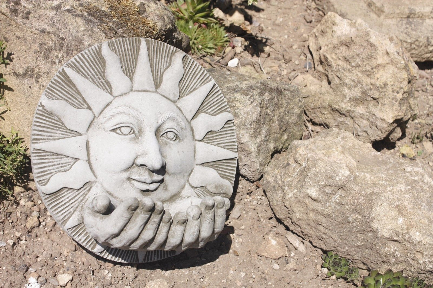 Handmade Stone Sun Garden Bird Feeder resting on rocky ground, showcasing detailed facial carving and food placement hands.