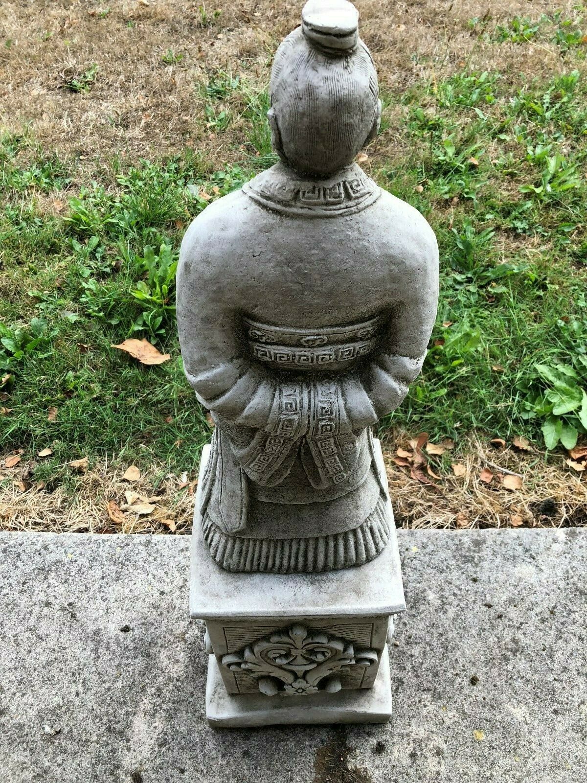 Back view of the Stunning Stone Zhan Shi Warrior Garden Plinth Statue with detailed robe patterns, placed on a concrete patio near grass.