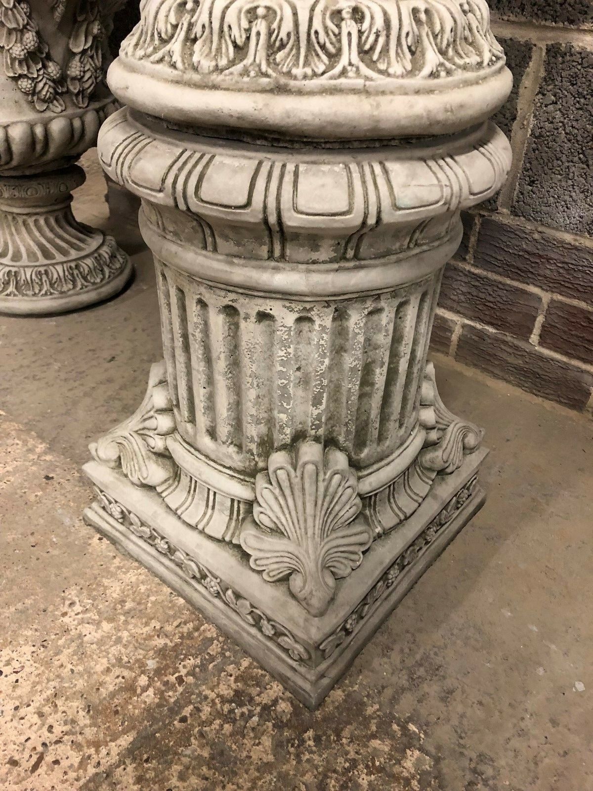 Close-up of ornate stone plinth base with fluted column and shell accents, supporting a Bacchus urn planter for outdoor decor.
