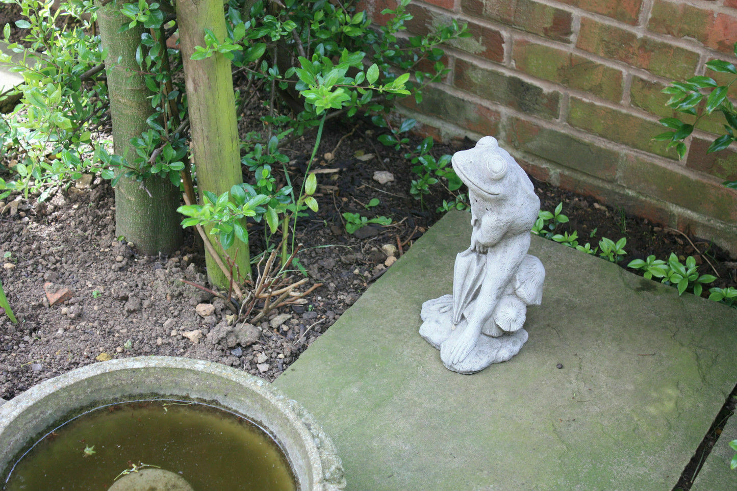 Stone Frog Garden Ornament with Umbrella placed beside a small pond and brick wall, enhancing garden ambiance with artistic flair.