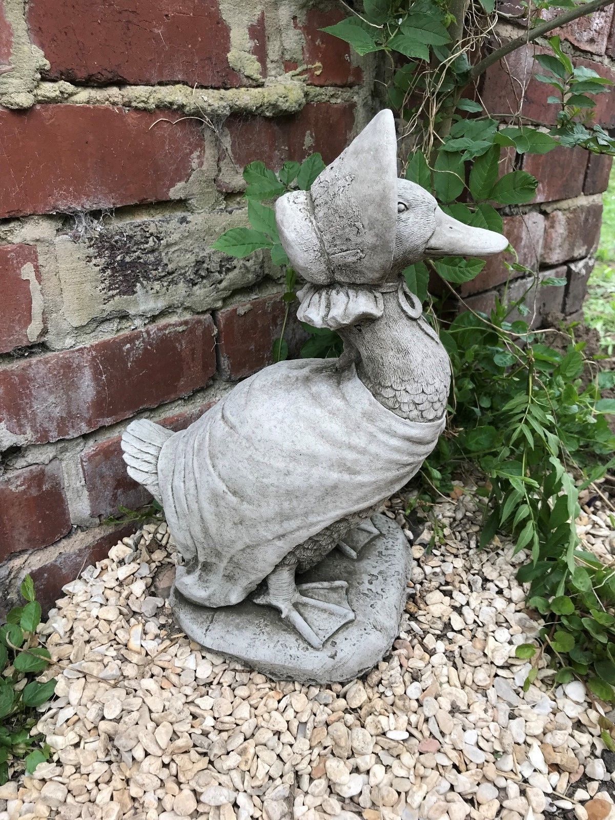 Side angle of stone duck ornament on gravel near brick wall. Weatherproof and ideal for enhancing garden aesthetics.
