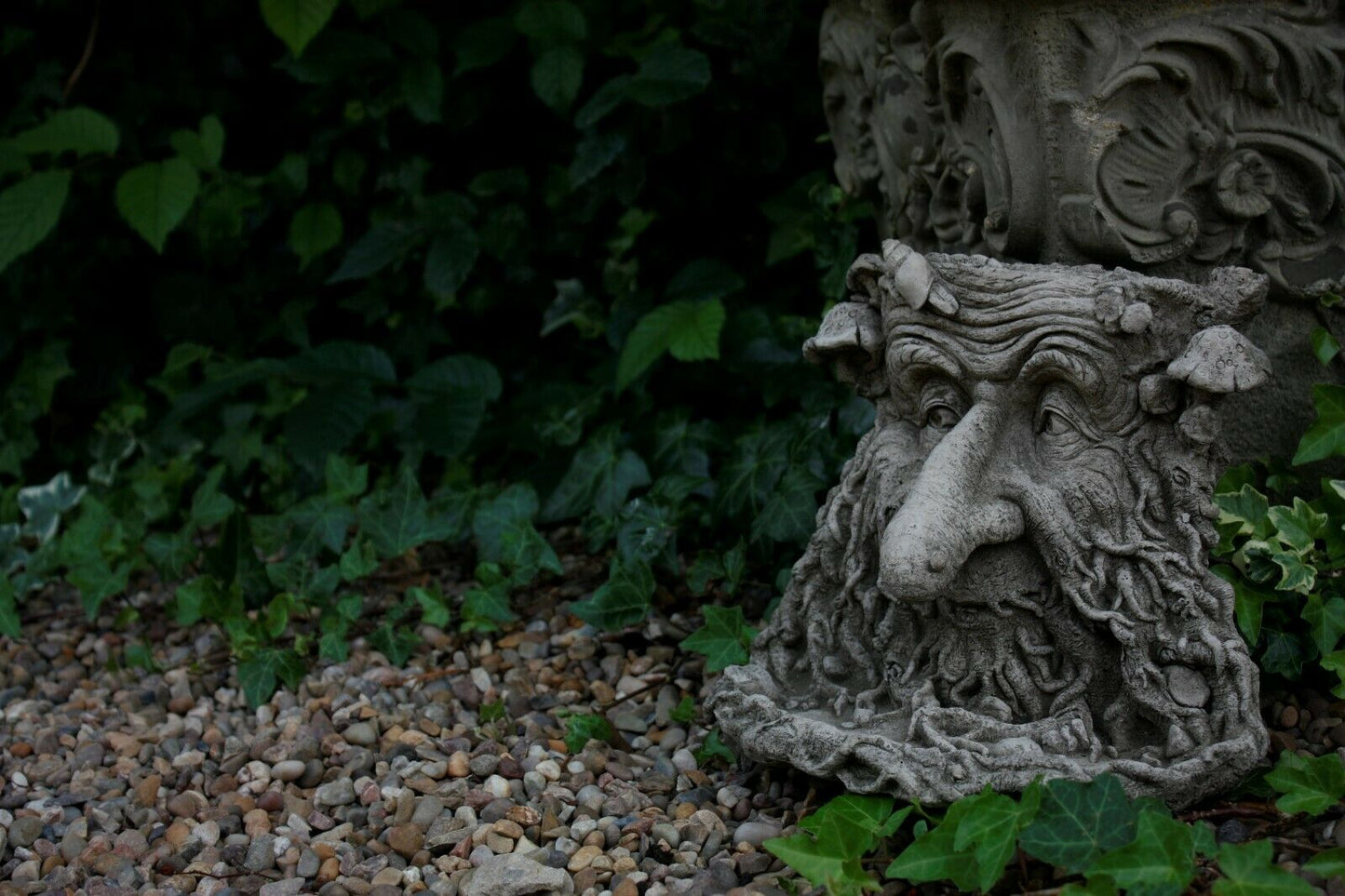 Detailed Stone Greenman Bird Feeder with long nose resting on gravel and ivy, offering a weather-resistant and artistic garden feature.