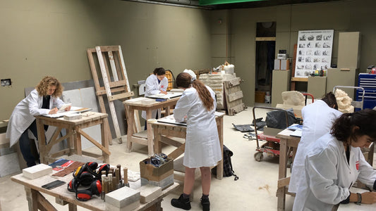 Aspiring sculptors in white coats practice stone carving in artisan workshop.