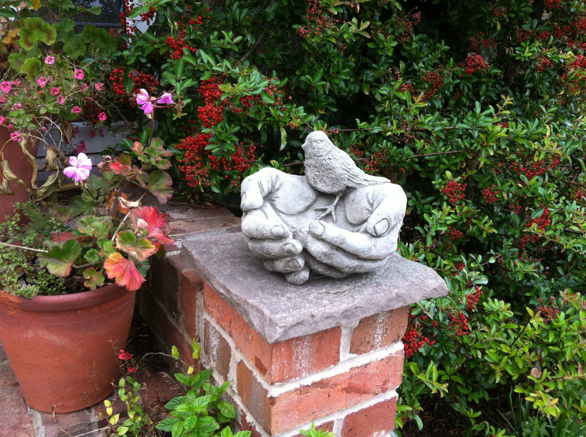 Decorative Bird Bath/Feeder in Stone Hands Design on a garden pillar, surrounded by vibrant plants, ideal for outdoor wildlife decor.