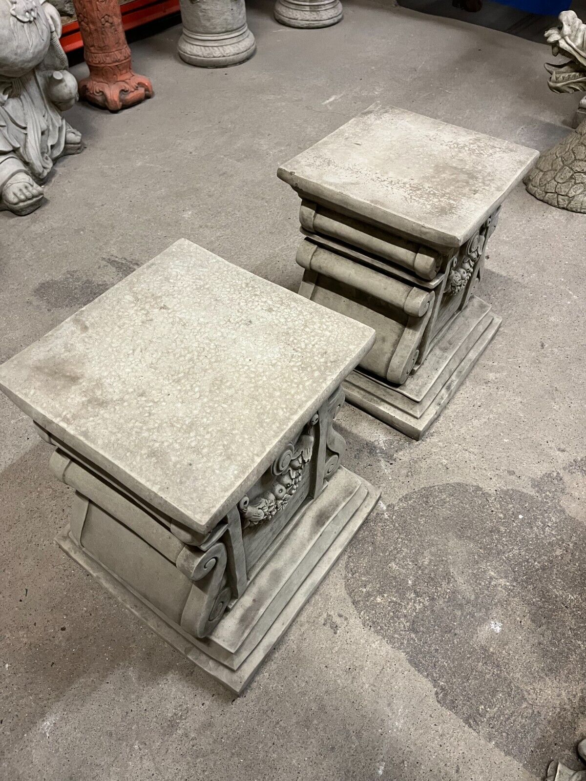 Angled view of antique-style garden plinths with scroll and garland motifs, crafted in heavy grey stone for outdoor use.