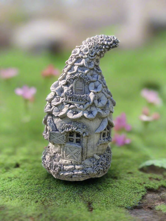 Stone Fairy Cottage garden ornament with floral roof detail, placed on mossy ground among pink flowers. Weatherproof and durable for outdoor decor.