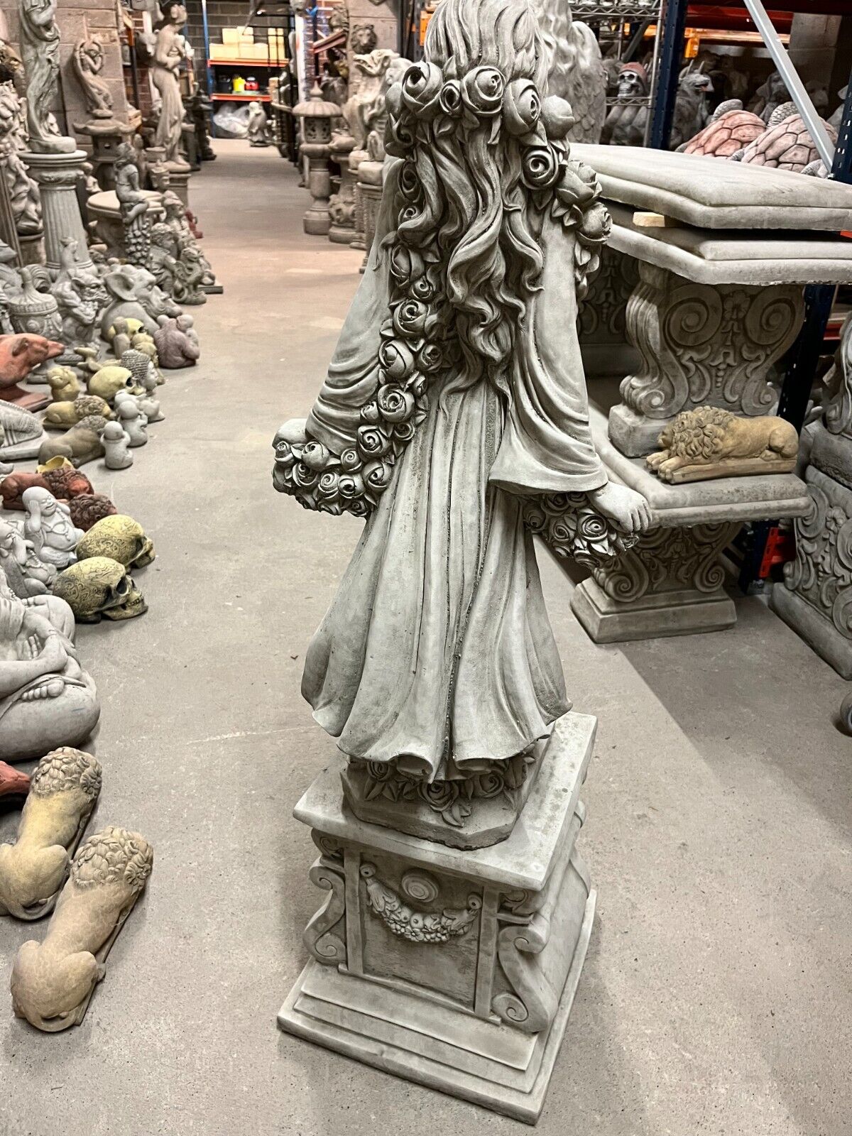 Rear view of antique finish garden statue with long flowing hair adorned with roses, standing on a decorative plinth.