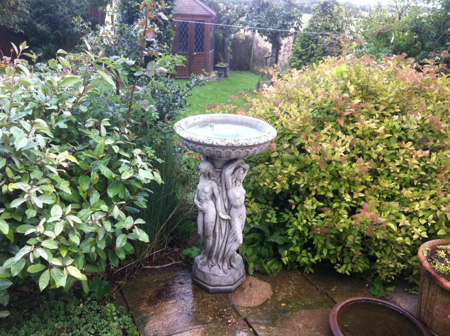 Large antique grey stone bird bath featuring Three Graces sculpture and raised perch, perfect for elegant garden settings.