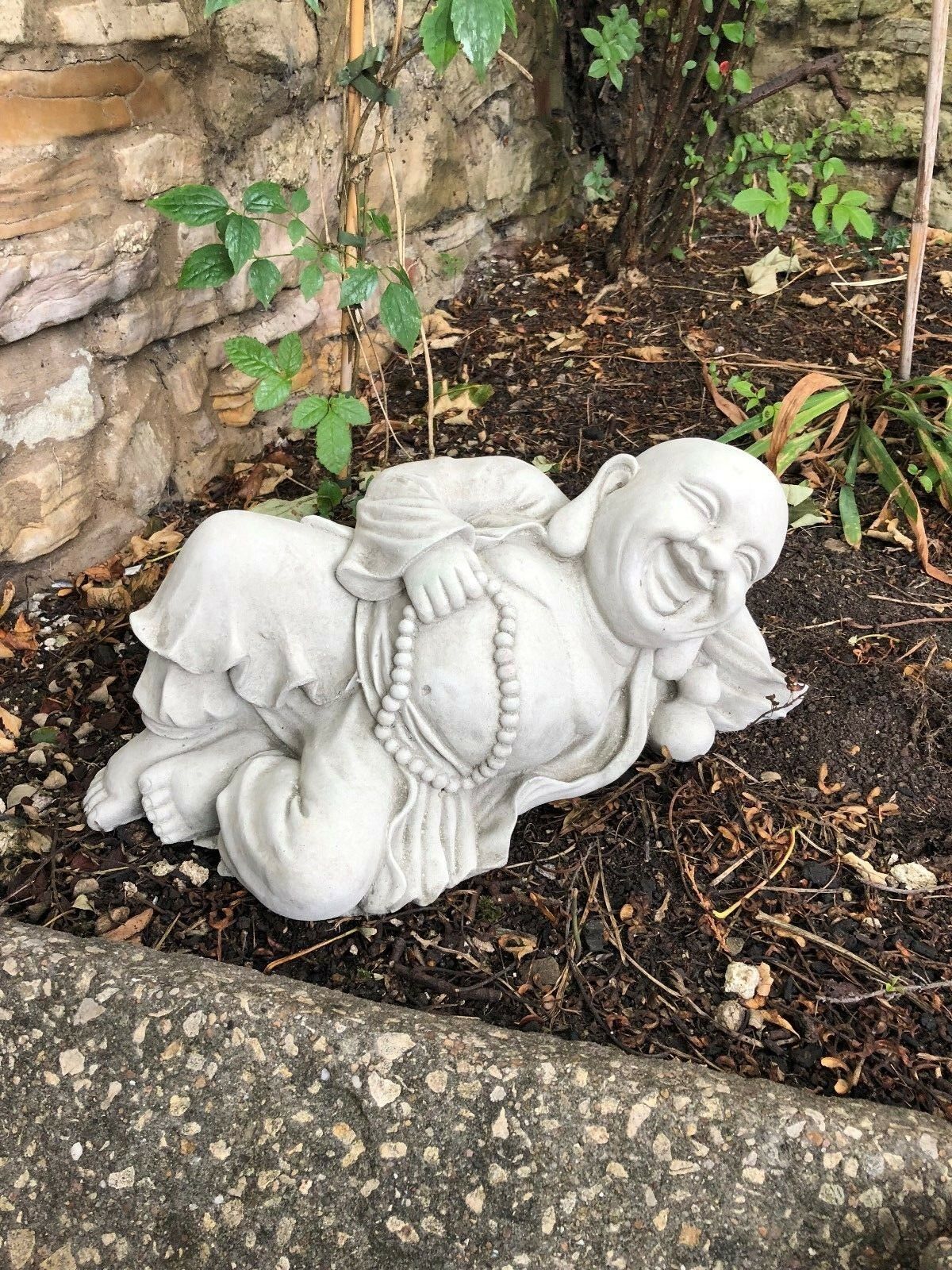 Stone Buddha sculpture lying on side with serene smile and prayer beads, enhancing garden tranquility and spiritual ambiance.