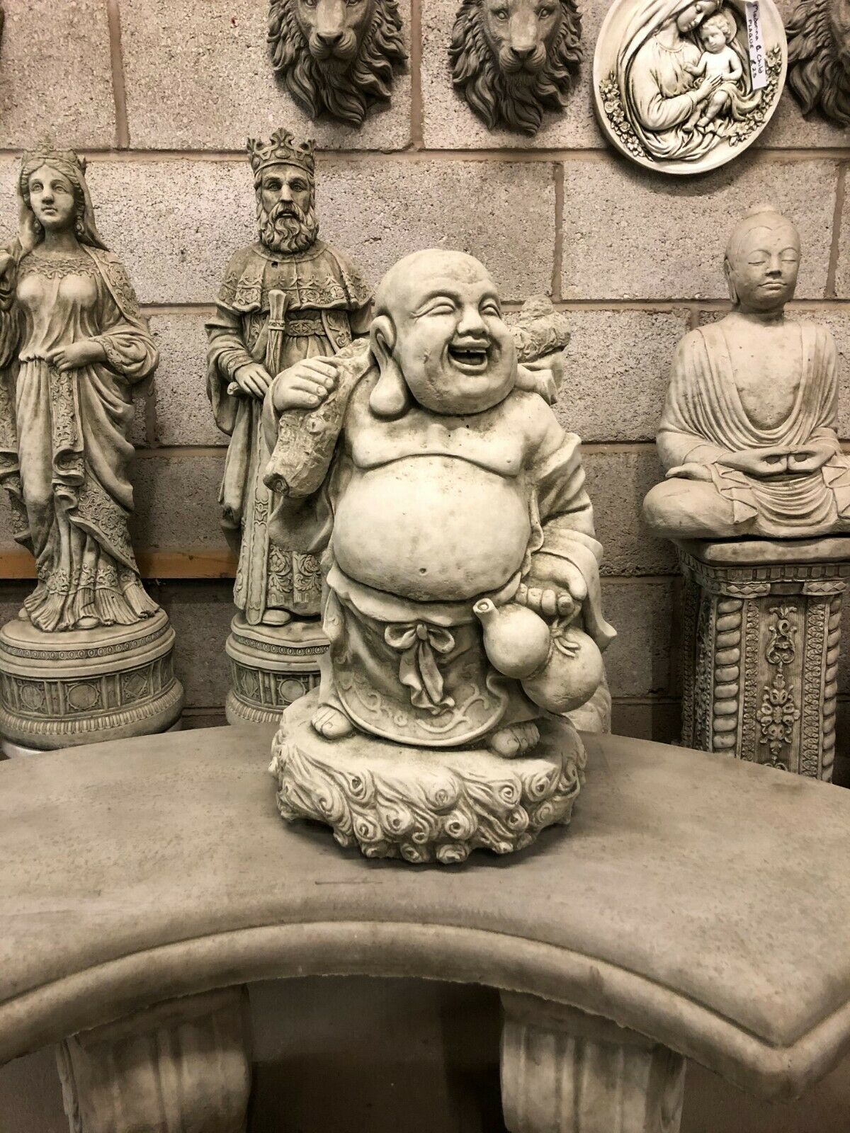 Stone Laughing Buddha Statue - Weather Resistant | Antique Finish, placed on a curved stone bench with detailed carvings, ideal for garden decor.