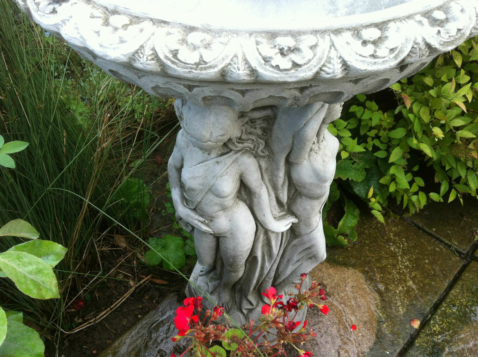 Ornate stone bird bath with mythological Three Graces figures and floral detailing, surrounded by blooming garden plants.