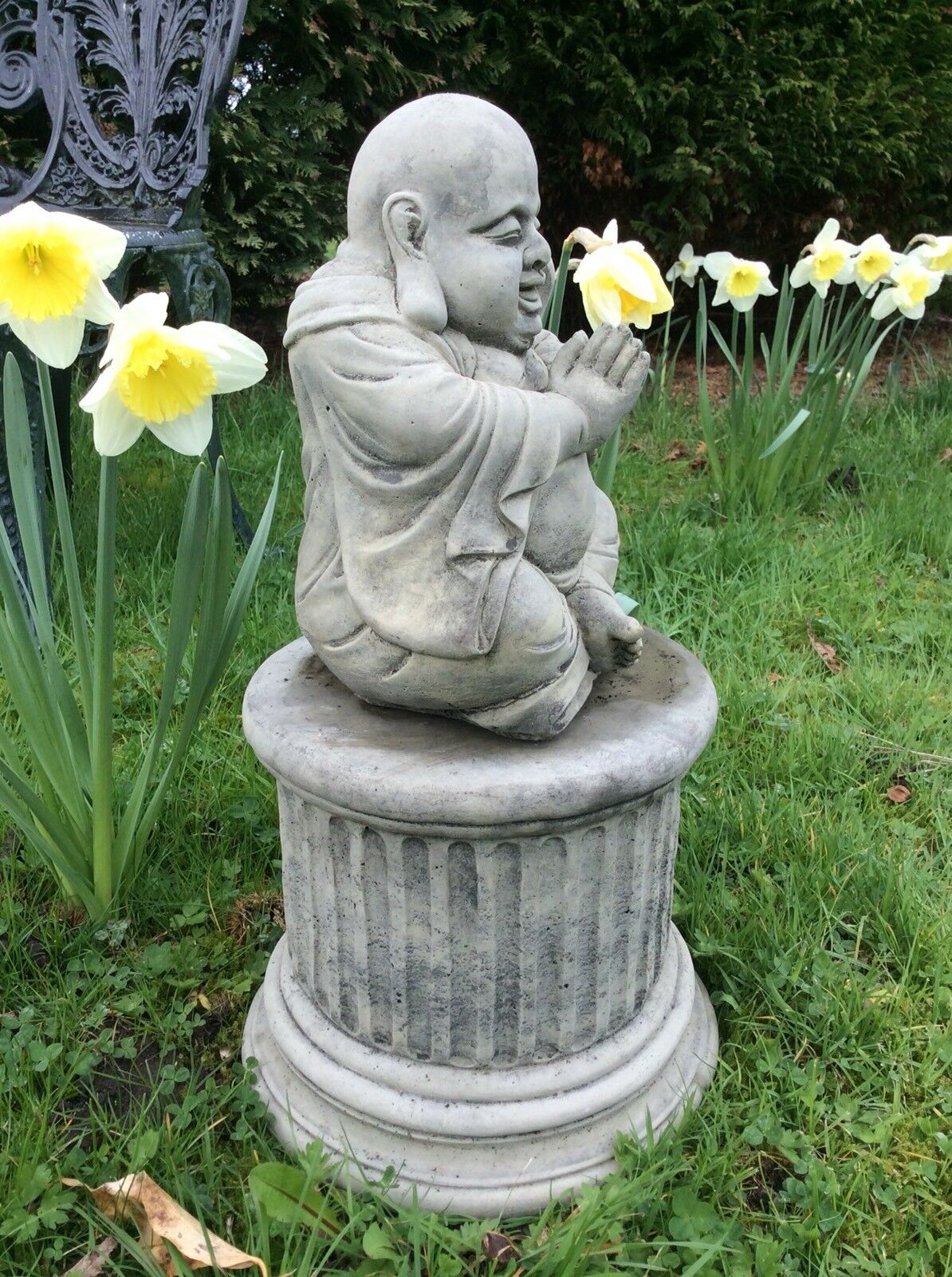 Side view of stone Happy Praying Buddha Statue on a fluted pedestal, perfect for enhancing garden serenity with durable outdoor decor.
