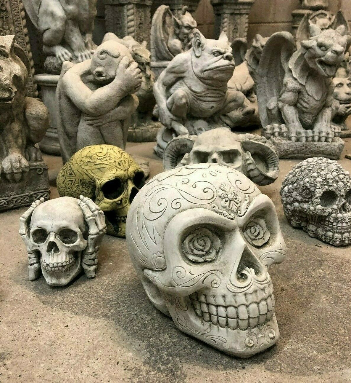 Antique Finish Stone Skull Garden Ornament among gothic statues. Features rose eyes and ornate carvings, perfect for themed garden decoration.