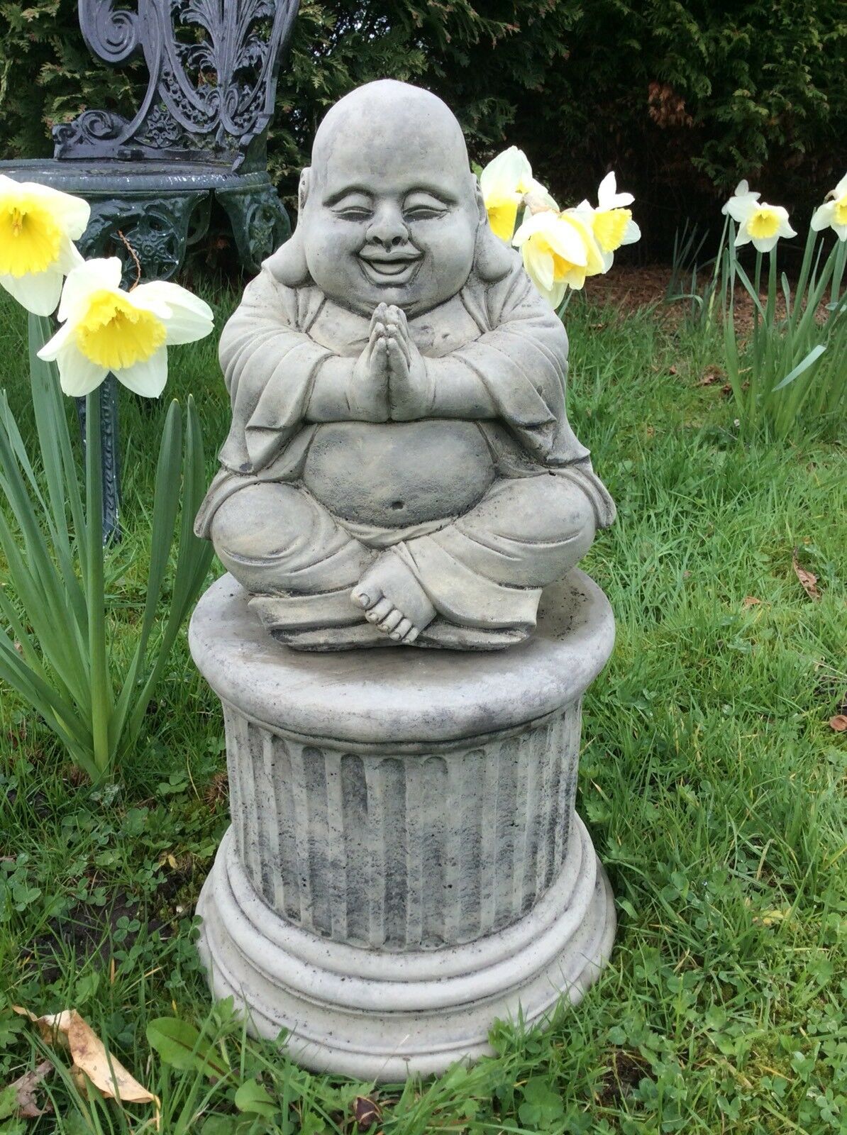 Front view of smiling stone Buddha statue in prayer pose, seated on a round pedestal, designed for tranquil garden decoration and lasting beauty.