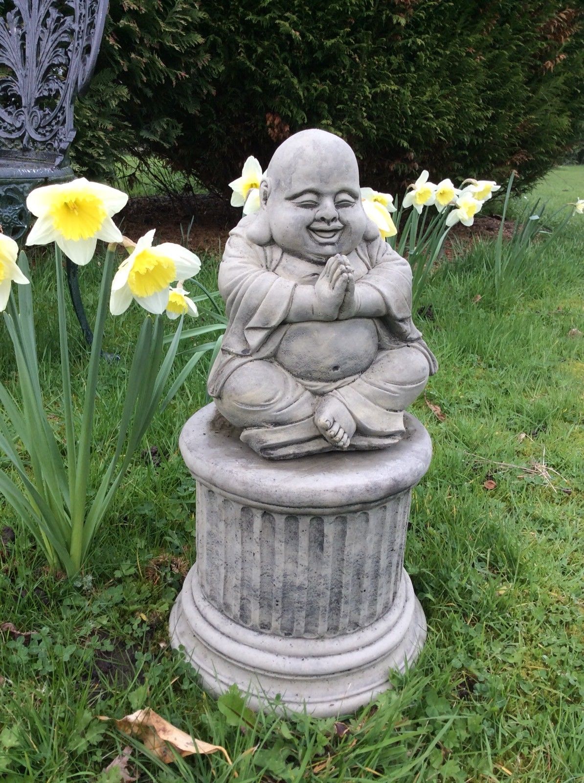 Angled view of Happy Praying Buddha Statue in stone, seated cross-legged on a plinth, ideal for peaceful and decorative garden settings.