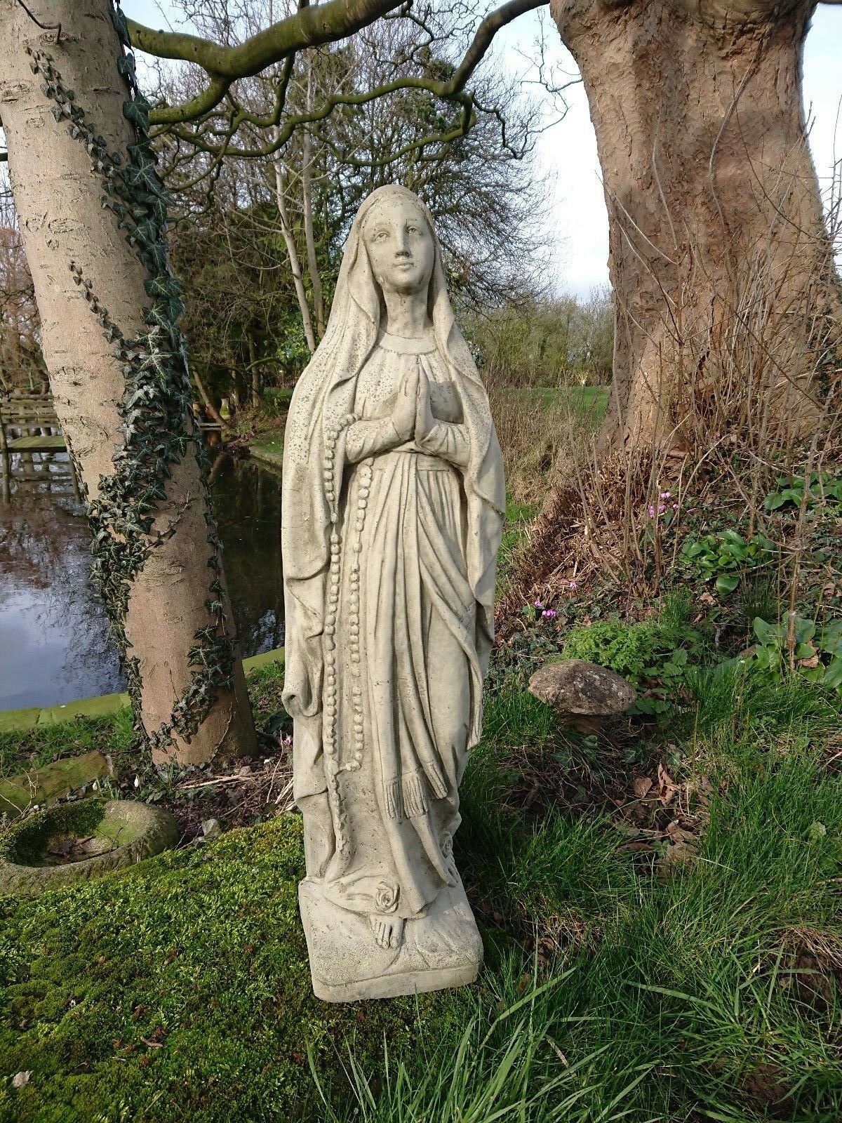 Stone Virgin Mary garden statue with antique finish, standing on mossy ground near a pond. Ideal religious decor for outdoor spaces.