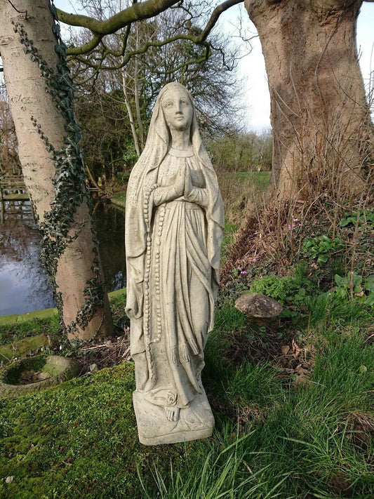 Stone Virgin Mary garden statue with antique finish, standing on mossy ground near a pond. Ideal religious decor for outdoor spaces.