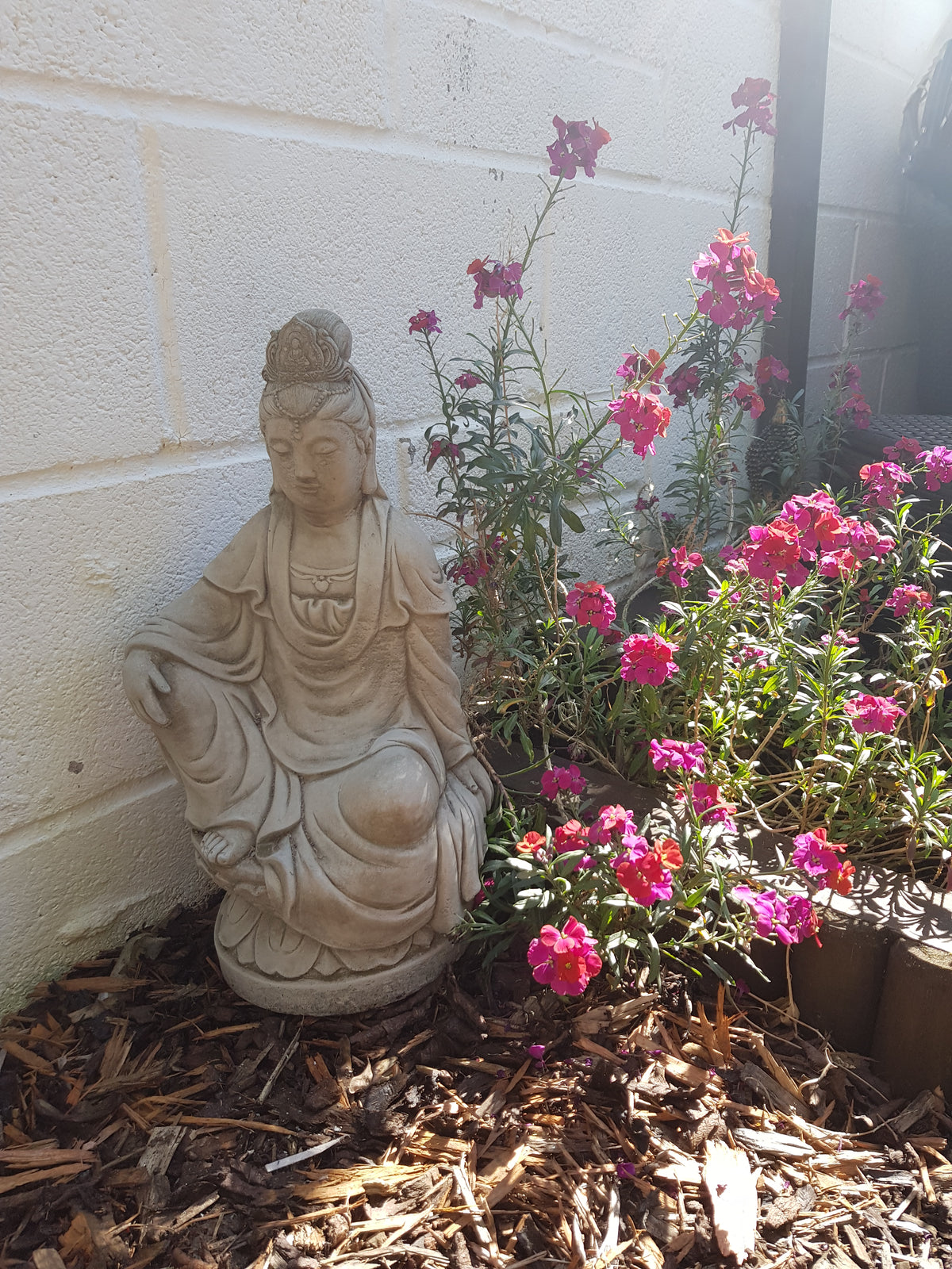 Kuan Yin garden statue placed among pink flowers and mulch. 18" tall stone sculpture adds spiritual charm to outdoor floral settings.