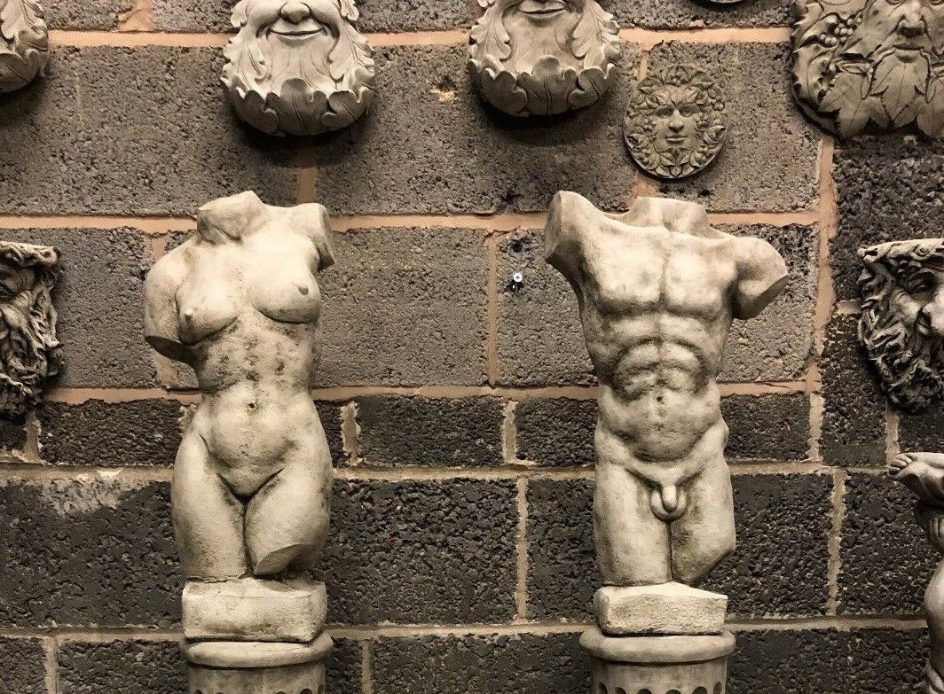 Side-by-side male and female torso statues on Grecian plinths. Antique grey limestone sculptures for classical garden aesthetics.