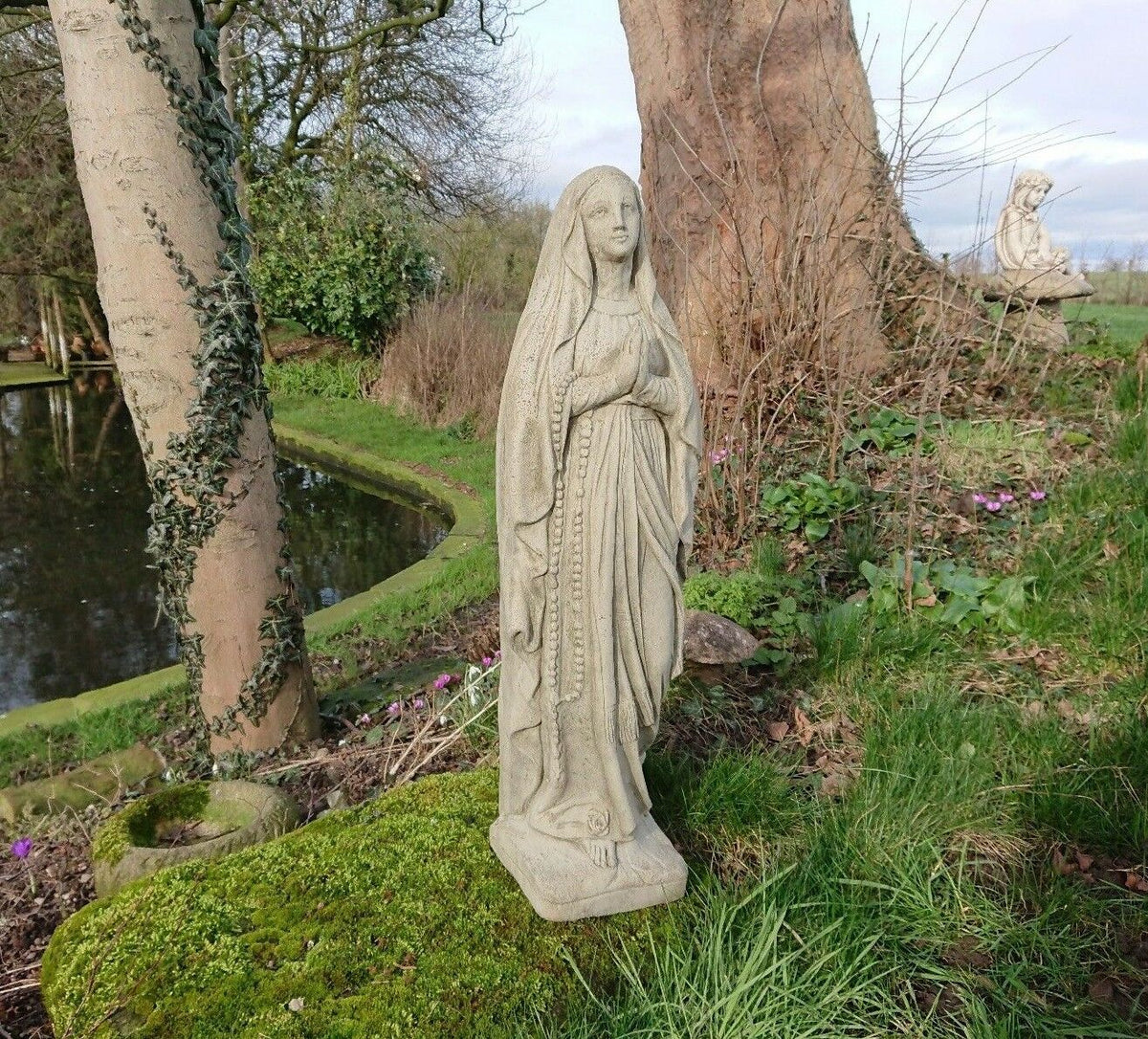 Durable limestone Virgin Mary statue in antique grey, placed in a garden by a pond. Perfect for Christian lawn ornamentation.