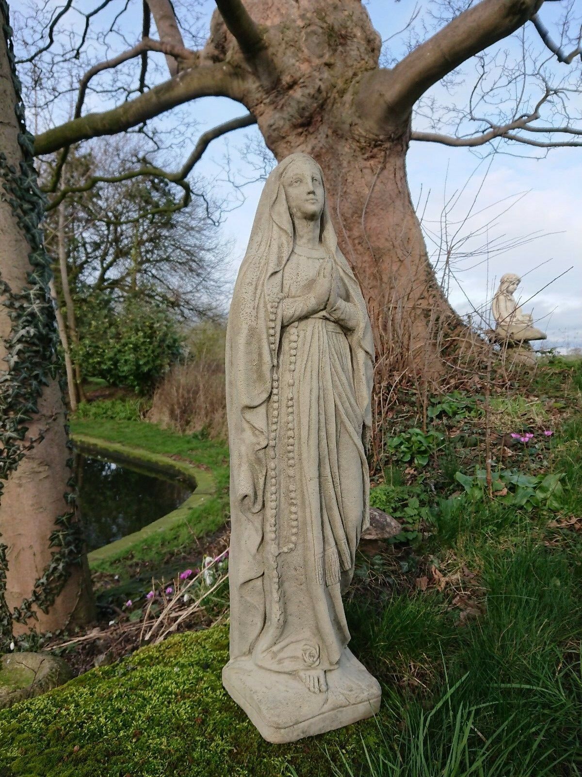 Detailed stone Virgin Mary garden sculpture with rosary, set in a natural outdoor setting. Weather-resistant religious garden decor.