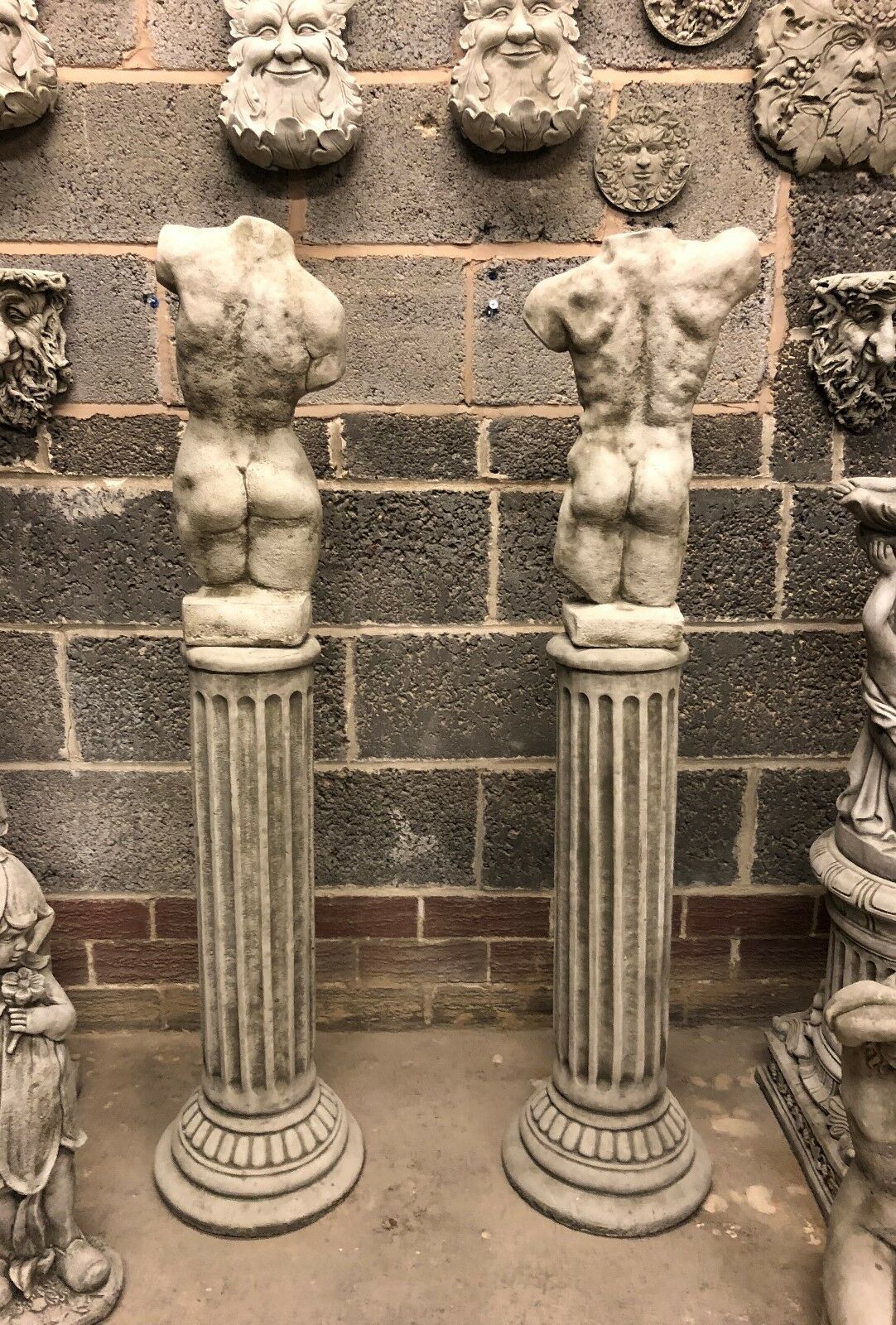 Rear view of stone torso statues on Grecian plinths, showcasing sculpted back and gluteal detail. Durable garden sculptures with classic design.