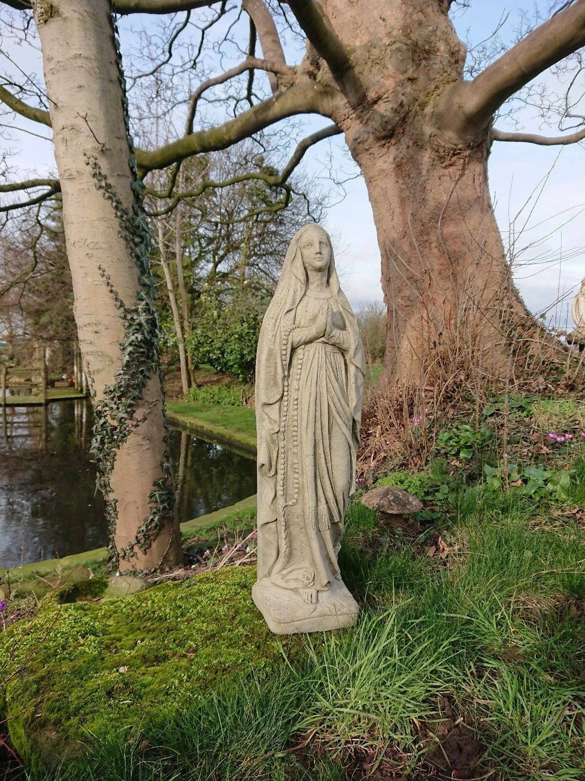 Handcrafted Virgin Mary garden statue in limestone with antique finish, placed near trees and a pond. Ideal for memorial or patio decor.