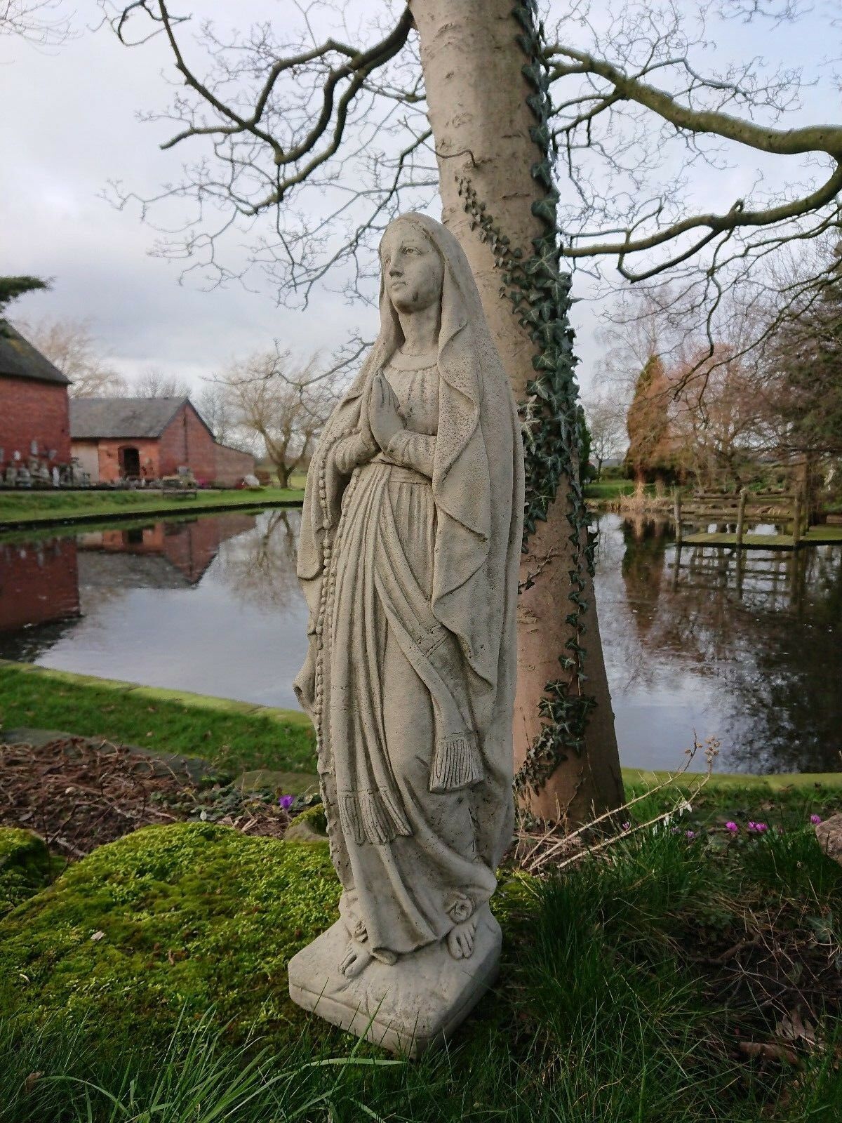 Outdoor Virgin Mary statue in antique grey stone, positioned beside a tree and pond. Durable religious ornament for garden landscapes.