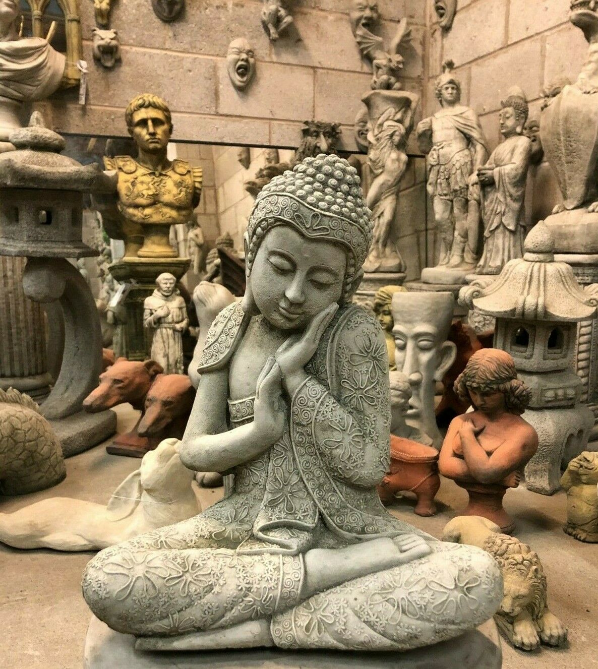 Stone Buddha Garden Ornament - Hands on Cheek in concrete finish, seated in meditation pose among various garden sculptures. Ideal for outdoor decor.