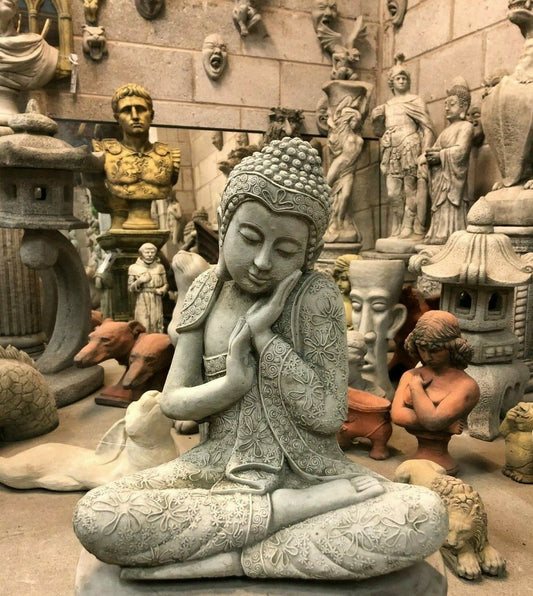 Stone Buddha Garden Ornament - Hands on Cheek in concrete finish, seated in meditation pose among various garden sculptures. Ideal for outdoor decor.