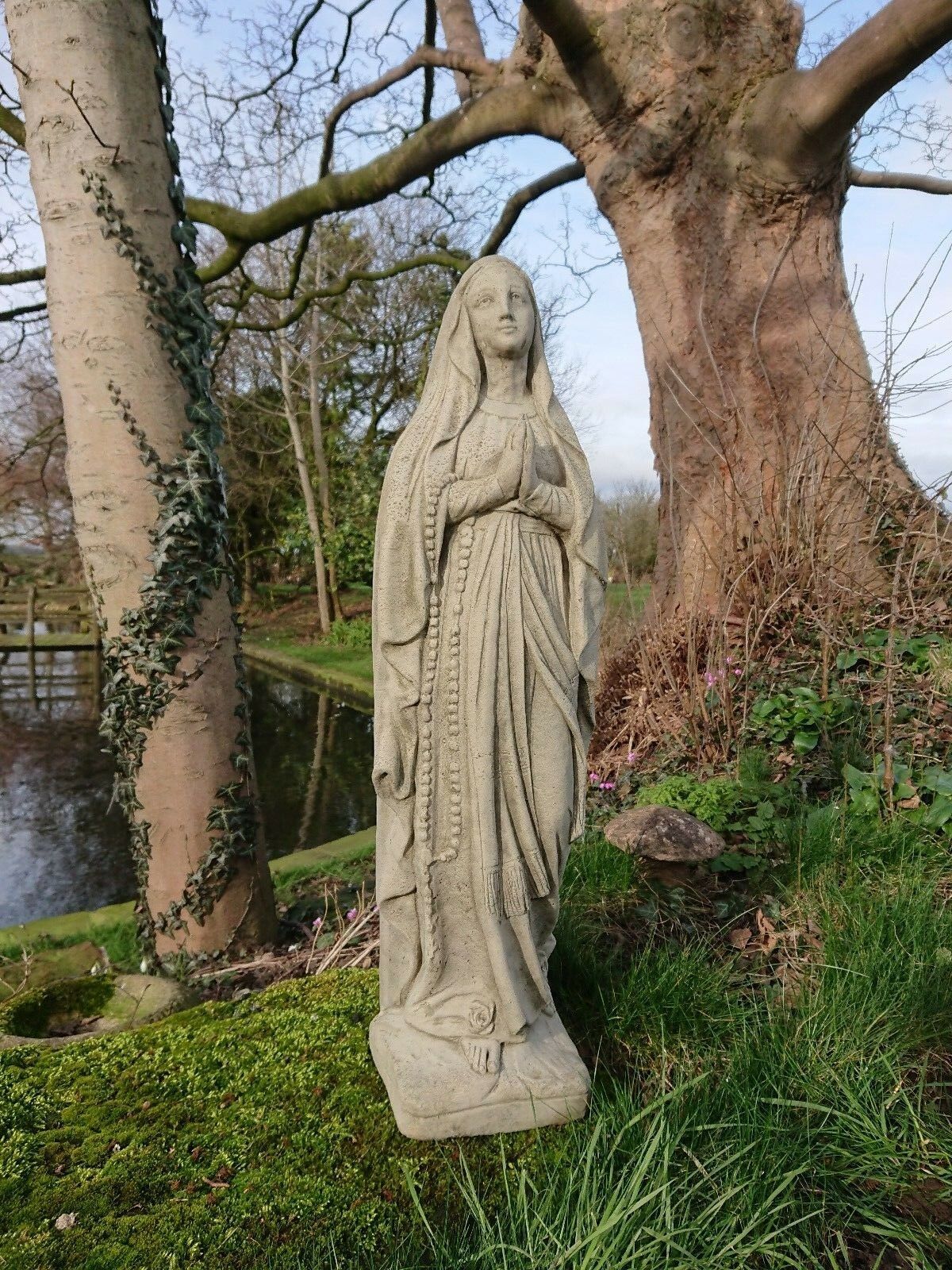 Solid stone Virgin Mary statue with detailed robe and rosary, set in a lush garden. Classic Christian decor for outdoor use.