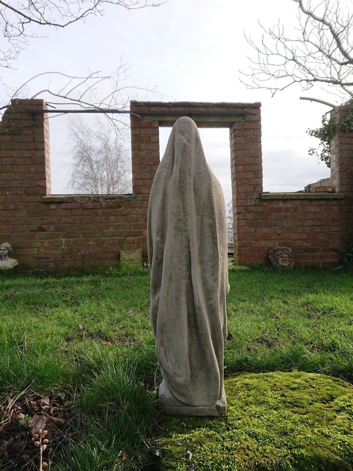 Back view of antique-finish Virgin Mary garden statue in limestone, facing a rustic brick structure. Suitable for religious garden themes.