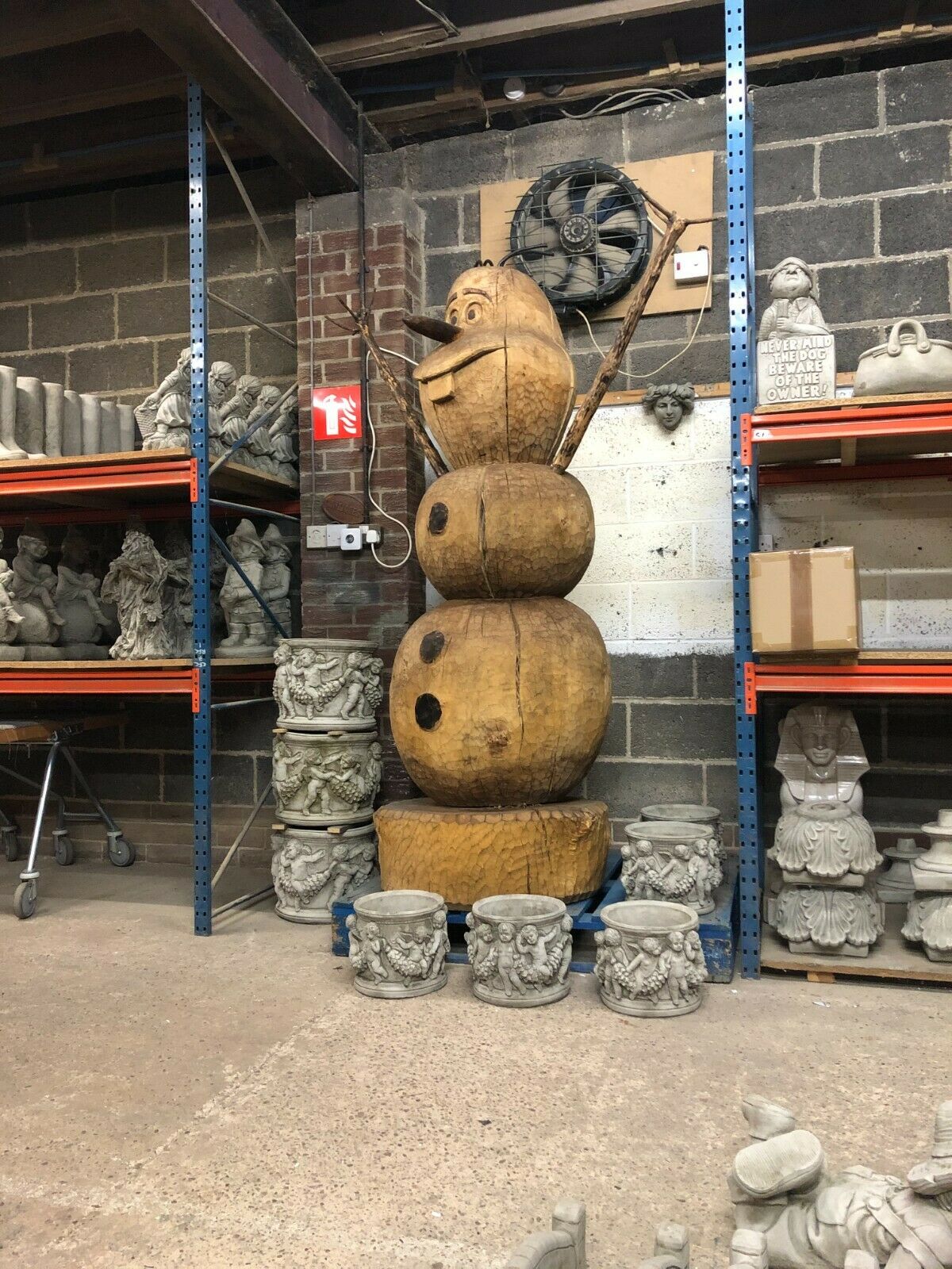 Full view of the Giant Wooden Olaf Garden Statue in a warehouse, surrounded by decorative stone planters and sculptures.