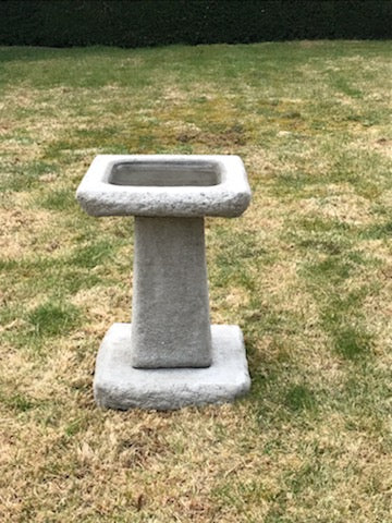 Stone bird bath with chunky square design placed on a lawn. Heavy and weather-resistant, ideal for garden or backyard decor.