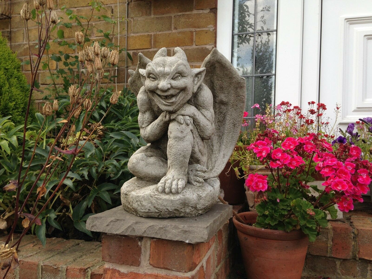 Winged Gargoyle Garden Ornament with stone finish placed on a brick pedestal among potted flowers and greenery. Ideal for outdoor gothic garden decor.