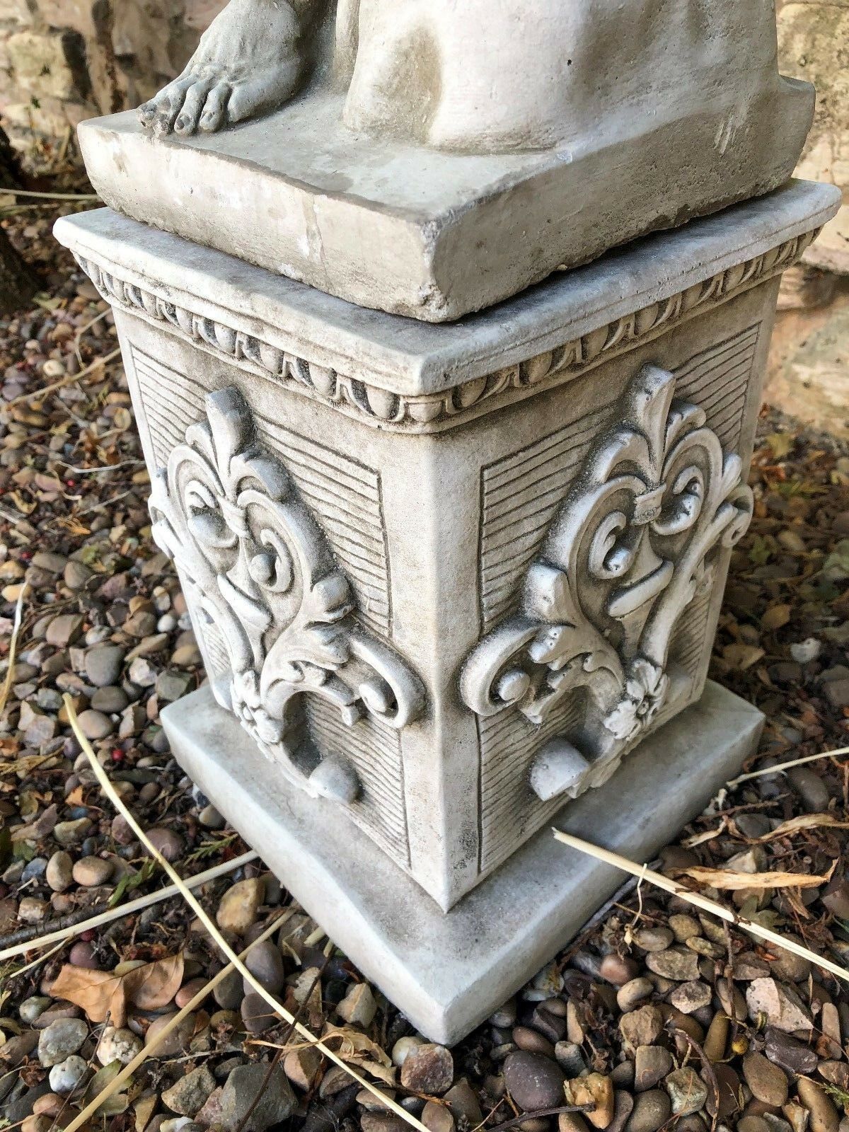 Detailed stone garden plinth with ornate scrollwork and flower motif, supporting a statue on a gravel patio. Heavy and weather-resistant.