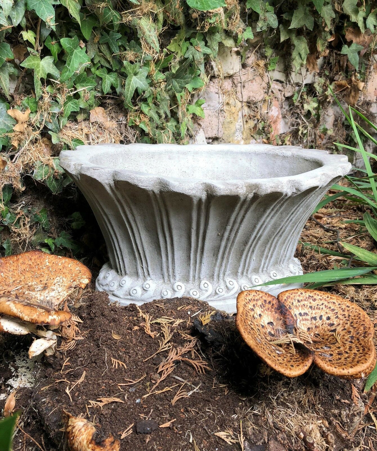 Fluted garden pot planter with stone finish placed in a garden bed, featuring elegant scalloped edges and durable limestone texture.