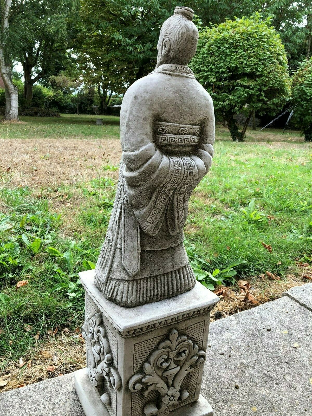 Rear view of the Zhan Shi Warrior Garden Statue showing detailed robe folds and hand placement, set against a lush garden background.
