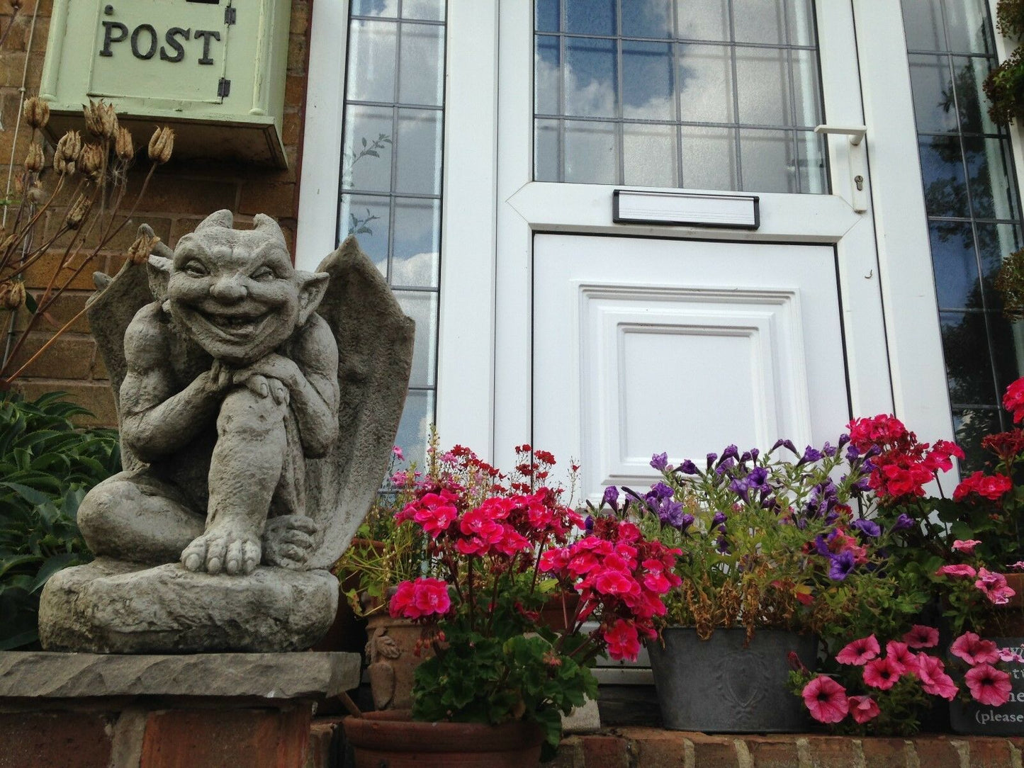 Smiling Winged Gargoyle Garden Ornament in front of a white door surrounded by colorful flowers, perfect for outdoor decoration and spiritual protection.