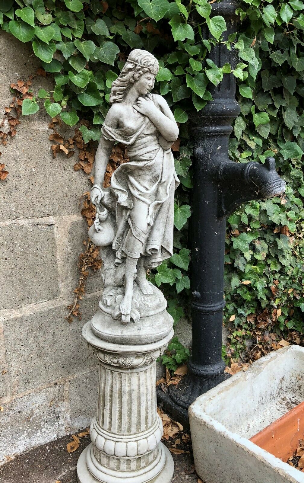 Garden Stone Statue - Jug Lady Plinth in antique grey limestone finish, standing beside a vintage black water pump, ideal for outdoor decor.