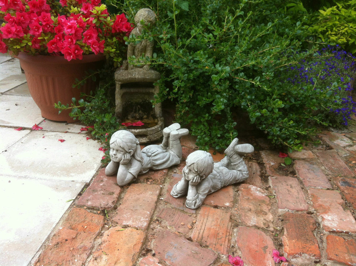 Stone garden ornaments - boy and girl laying pair on rustic brick patio surrounded by lush greenery and flowers. Durable limestone sculptures with antique finish.