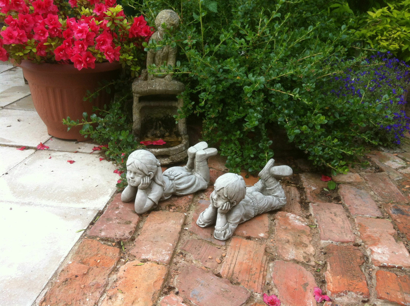 Stone garden ornaments - boy and girl laying pair on rustic brick patio surrounded by lush greenery and flowers. Durable limestone sculptures with antique finish.