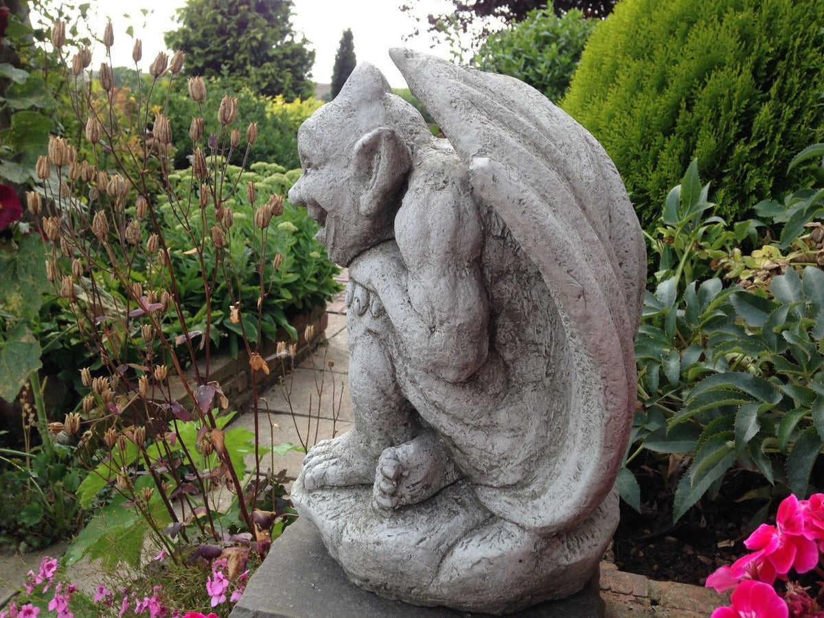 Profile view of Winged Gargoyle Garden Ornament with intricate stone detailing, set in a landscaped garden with flowering plants and shrubs.